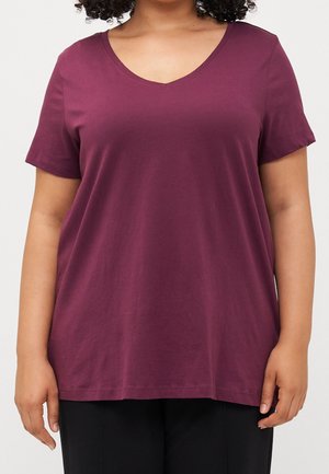 Frau trägt ein taubenblaues Kurzarm-T-Shirt mit V-Ausschnitt und schwarze Hose, Bildausschnitt von der Schulter bis zur Taille.