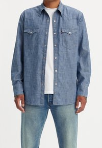 Camicia western in denim azzurro chiaro con bottoni a pressione, yoke appuntito, due tasche sul petto e maniche lunghe. Indossata sopra una t-shirt bianca e jeans.