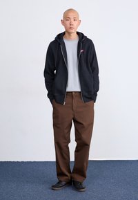 Juoda užtrauktuko megztinis su pilku marškinėliu, derinamas su rudomis kelnėmis. Modelis dėvi juodus batus. Paprastas dizainas, laisvas pjūvis ir švelnios tekstūros.