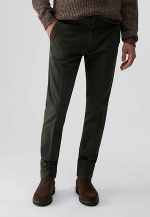 Homme portant un pantalon vert foncé, des bottes en daim marron et un pull tricoté marron avec une main dans la poche.