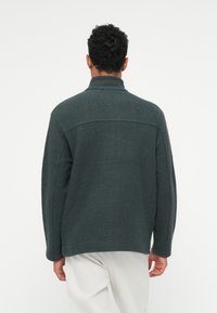 Pullover de polar verde escuro com um colarinho alto, ajuste descontraído e detalhes de costura visíveis; superfície texturizada, usado sobre calças de tom claro.