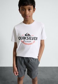 Bílý bavlněný tričko s černým a červeným logem Quiksilver, spárované se šedými kraťasy na šňůrku. Jednoduchý, ležérní design bez vzorů.