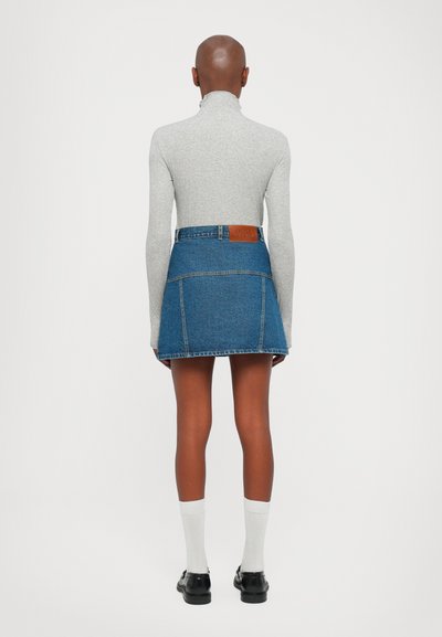 Denim mini svārki ar strukturētu dizainu, zila krāsa, redzamās šuves un marķēts ādas plāksteris jostasvietā. Pelēks augsta kakla krekls un baltas zeķes.