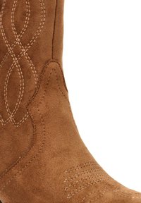 Bota vaquera de ante marrón con patrones de costura intrincados, punta afilada y superficie texturizada. Los acentos incluyen costuras decorativas.