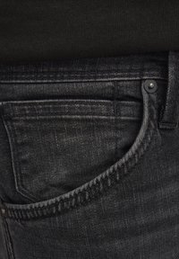 Vaqueros de mezclilla negra con una textura sutil, que cuentan con un bolsillo frontal y un botón de metal. La costura es visible a lo largo de los bordes del bolsillo.