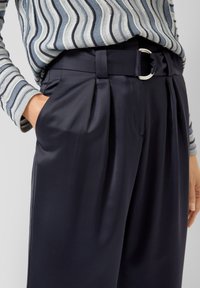 Marineblauwe geplooide broek met een gladde textuur en een stoffen riem met een zilveren ronde gesp. Gecombineerd met een gestreept shirt met lange mouwen.