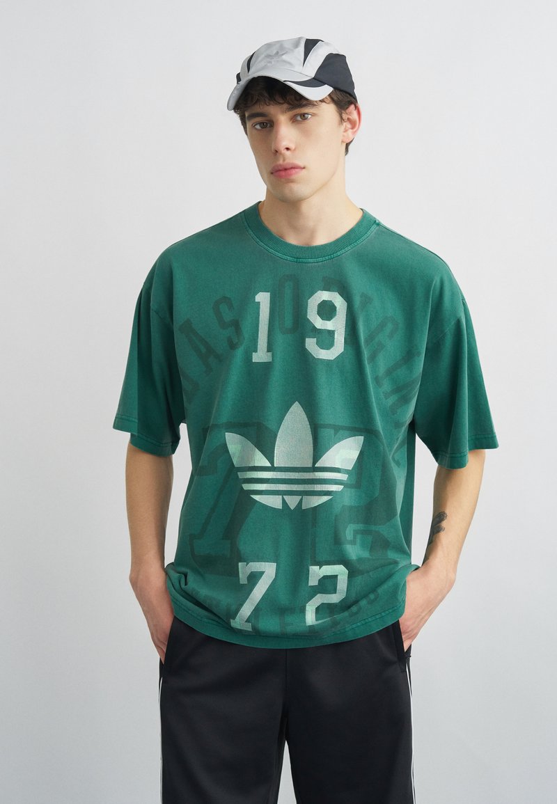 Jeune homme portant un t-shirt Adidas vert avec "1972" et une casquette noir et blanc, debout les mains dans les poches devant un fond uni.
