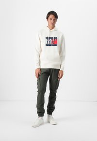 Krémová mikina s kapucí a námořnicky modrým a červeným logem 'Tommy Jeans', v kombinaci s olivově zelenými cargos kalhotami. Model má na sobě bílé sneakers. Jednoduchý, moderní střih.
