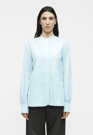 Γυναίκα με μακριά σκούρα μαλλιά που φορά ένα γαλάζιο πουκάμισο με κουμπιά και μανταρινένιο γιακά και μαύρο παντελόνι, στέκεται μπροστά σε απλό φόντο.