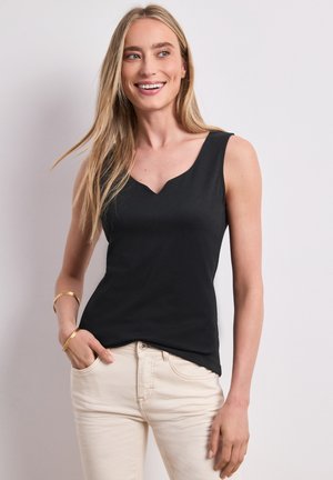 Femme souriante aux longs cheveux blonds portant un haut noir sans manches, un pantalon beige et des bracelets dorés, debout devant un fond clair.