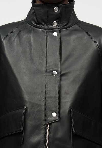 Κοντινή λήψη μπροστινού μέρους μαύρου δερμάτινου μπουφάν με ψηλό γιακά, κουμπιά πατήματος, φερμουάρ και τσέπες με καπάκι σε κάθε πλευρά.