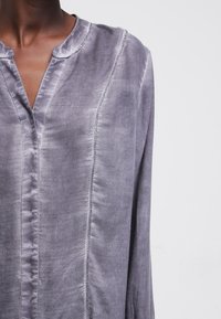 Blusa grigia a maniche lunghe realizzata in tessuto liscio con scollatura a V e una sottile cucitura decorativa lungo la parte anteriore. Design minimalista.