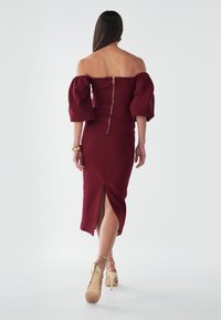 Abito borgogna off-the-shoulder con maniche a sbuffo, design aderente, zip posteriore e spacco laterale. Modella che indossa sandali con tacco alto e fasce.