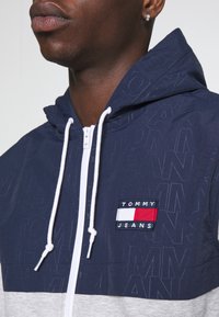 Chaqueta con capucha de color navy con un diseño texturizado. Presenta una sección inferior gris, cordones blancos y un parche de logo multicolor prominente.