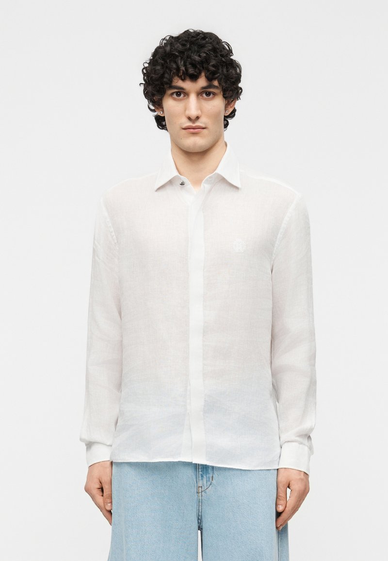 Άνδρας με σγουρά μαλλιά που φοράει ημιδιαφανές λευκό πουκάμισο με κουμπιά και ανοιχτόχρωμο τζιν σε απλό λευκό φόντο.