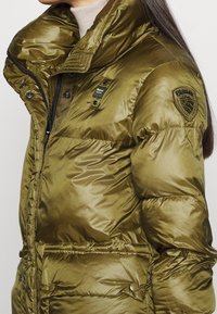 Giubbotto imbottito verde oliva con finitura lucida, colletto alto, chiusura con zip e tasche laterali. Presenta una patch con logo sulla spalla.