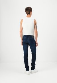 Fehér ujjatlan pólót viselő férfi, sötétkék farmernadrágban és fehér sportcipőben, háttal állva, bal karján tetoválással, sima fehér háttér előtt.