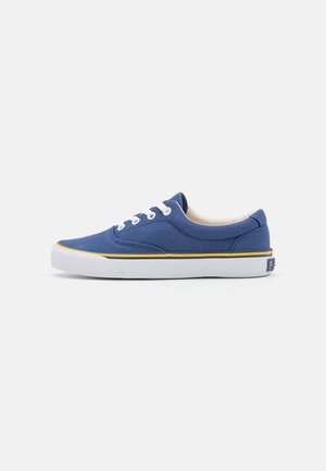 Singola sneaker in tela blu a taglio basso con lacci bianchi, suola bianca e sottile striscia gialla sopra la suola.
