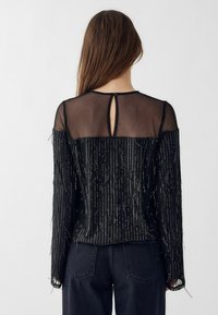 Czarna bluzka z długimi rękawami z przezroczystym tyłem i metalicznymi frędzlami. Zapięcie na guzik przy szyi, warstwowa tekstura i luźny krój.