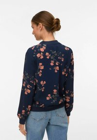 Kobieta z brązowym kokiem, ubrana w granatową, kwiecistą bluzkę z długim rękawem i jasnoniebieskie dżinsy, pokazana od tyłu na białym tle.