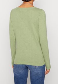 Maglione a maglia verde chiaro con un motivo testurizzato, maniche lunghe e scollatura rotonda. L'orlo inferiore è leggermente più lungo sul retro.