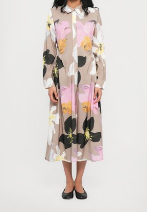 Femme portant une robe midi beige avec de grandes impressions florales roses, noires, blanches et jaunes et des ballerines noires, debout devant un fond blanc.