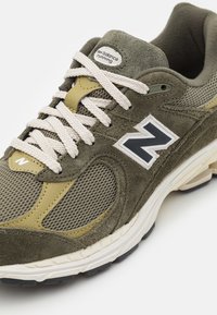 Olivgrön New Balance löparsko med mesh-paneler, mockaöverdrag, krämfärgade snören och vit "N"-logotyp på sidan, vinklad vy.