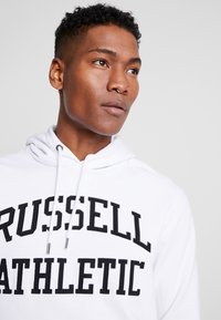 Sudadera blanca con el texto negro "RUSSELL ATHLETIC", cordones y un bolsillo frontal. Tejido suave de mezcla de algodón, con puños y dobladillo acanalados.