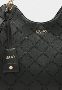 Sac à main en cuir noir texturé avec motif logo embossé, quincaillerie en ton doré et une étiquette amovible avec le nom de la marque.
