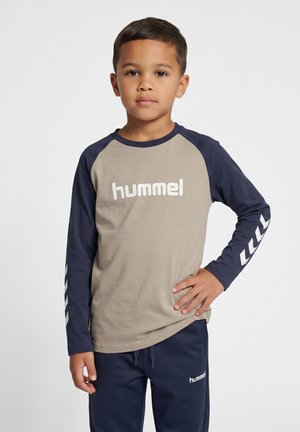 Dreng med kort mørkt hår står forfra iført en beige og marineblå langærmet skjorte og marineblå bukser med "hummel"-logo.