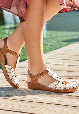 Sandalias de cuero marrón y blanco con cortes florales, usadas en pies cruzados de pie en un muelle de madera junto al agua.