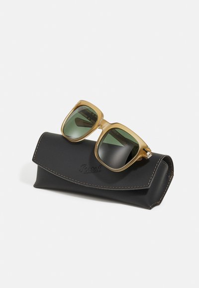 Óculos de sol de plástico castanho com lentes verdes, colocados numa capa de pele preta com costura castanha e um logotipo.