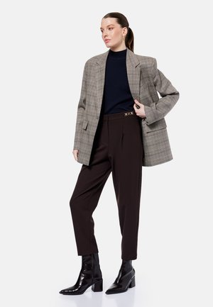 Vrouw draagt een grijs geruit blazer over een zwarte coltrui, donkere hoge taille broek met een gouden gesp en zwarte enkellaarsjes met hak, staand.