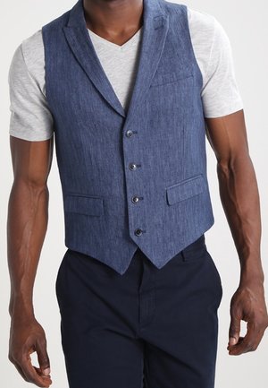 Uomo che indossa un gilet blu con bottoni sopra una maglietta grigia chiara a maniche corte con scollo a V e pantaloni scuri, in piedi davanti a uno sfondo neutro.