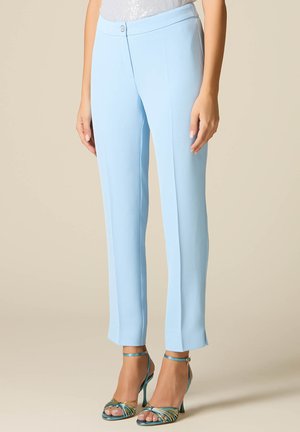 Žena v světle modrých kalhotách na míru a tyrkysově zelených sandálech na vysokém podpatku stojí před neutrálním pozadím.