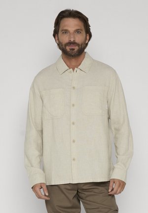 Mann mit Bart, der ein beiges Hemd mit Knöpfen und zwei Brusttaschen sowie braune Hosen trägt, steht vor einem schlichten weißen Hintergrund.