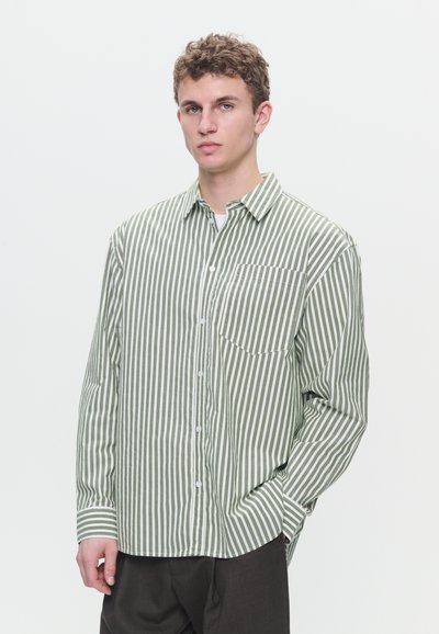 Young man with curly hair wearing green and white vertical striped button-up shirt and dark pants, standing with one hand in pocket.