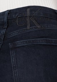 Dunkelblaue Jeans mit einem erhabenen schwarzen gestickten "CK"-Logo und dem Schriftzug "CALVIN KLEIN JEANS" am Bund; glatte Textur.