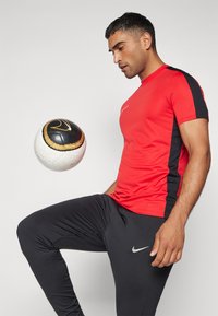 Rød atletisk T-shirt med sorte sidepaneler og et Nike-logo, parret med sorte tætsiddende bukser. En fodbold svæver nær personens fod.