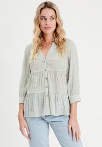 Blouse rayée vert clair avec un devant à boutons, un col en V et un design à étages. Présente des manches trois-quarts et un tissu doux et texturé.