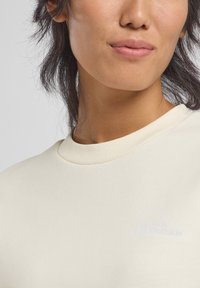 Camicia a maniche lunghe aderente di colore crema con collo a girocollo ribassato e un logo discreto sul petto. Texture liscia e morbida.