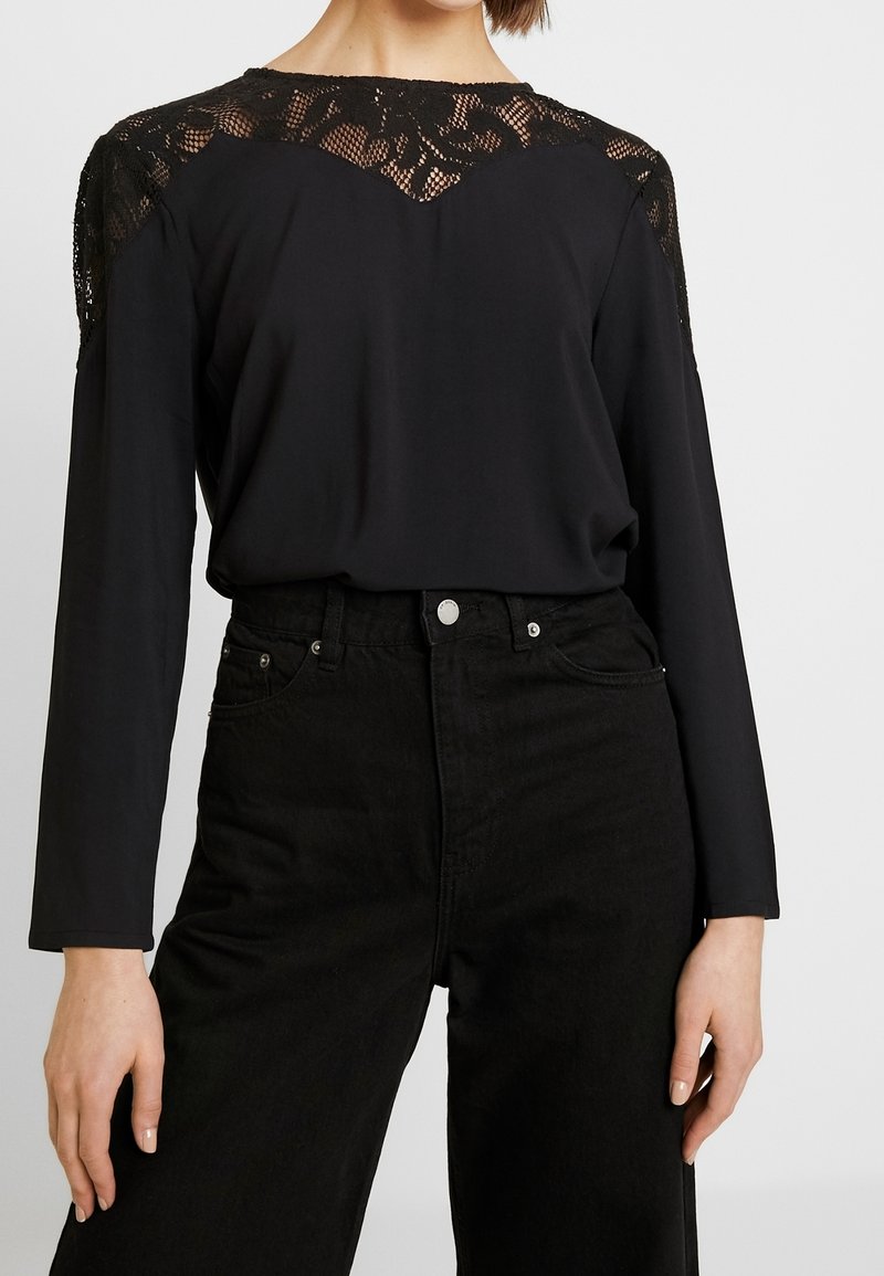 Kvinna som bär en svart långärmad topp med spetsdetaljer på axlarna och övre bröstet, tillsammans med högmidjade svarta jeans.