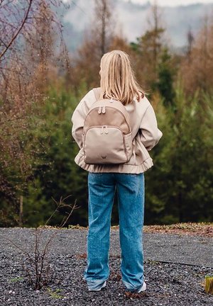 Beiger Rucksack mit runder Form, doppelten Reißverschlüssen und verstellbaren Trägern. Kombiniert mit einer leichten Jacke und blauen Jeans. Natürlicher Outdoor-Hintergrund.