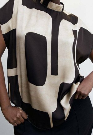 Kvinde iført en løs beige top med dristige, sorte abstrakte geometriske mønstre, kombineret med sorte bukser, hænderne hvilende på hofterne.