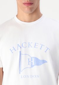 White cotton t-shirt featuring a light blue printed logo with a flag and text reading "HACKETT LONDON" in a classic font.