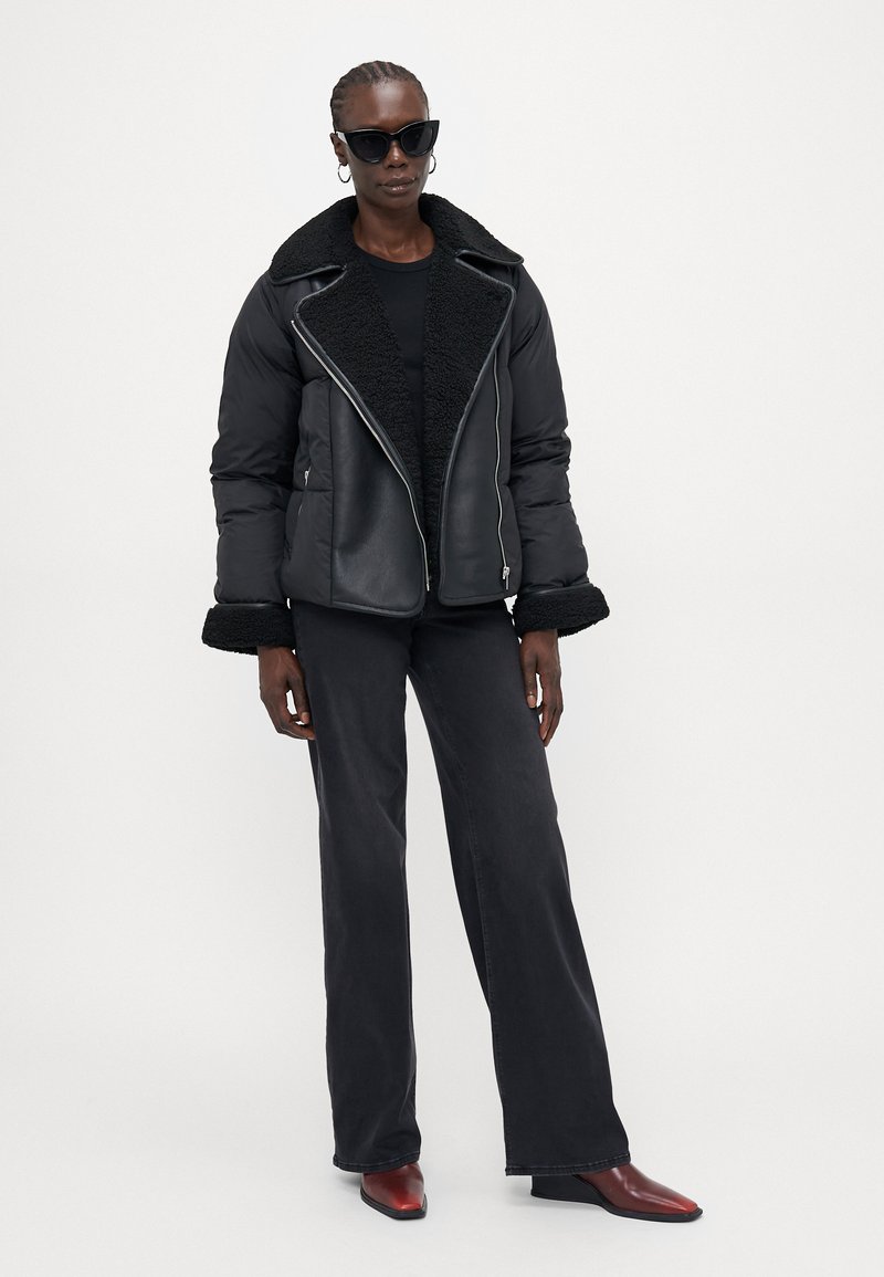 Giacca nera trapuntata con colletto in shearling, apertura con zip, abbinata a pantaloni neri a gamba larga e stivaletti rossi con punta a punta. Gli accessori includono occhiali da sole neri.