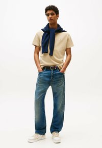 T-shirt beige con un piccolo logo, maglione navy appoggiato sulle spalle, jeans in denim blu, cintura nera e calzature bianche.