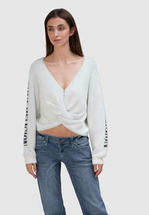 Weißer, flauschiger Pullover mit tiefem V-Ausschnitt, gedrehter Vorderseite und langen Ärmeln, die mit schwarzer Schrift an den Bündchen verziert sind. Kombiniert mit blauen Jeans.
