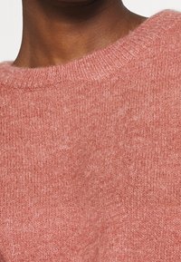 Roze gebreide trui met een ronde hals, zachte textuur en een iets fuzzy afwerking. Gedetailleerde stiksels zijn zichtbaar langs de halslijn.