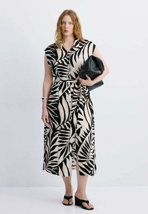 Femme aux cheveux roux bouclés portant une robe sans manches noir et blanc à motif feuilles, tenant une pochette noire et portant des sandales compensées noires.